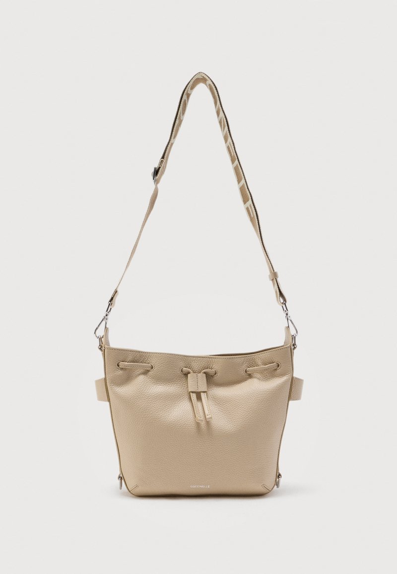 Μπεζ δερμάτινη τσάντα τύπου πουγκί με κλείσιμο κορδονιού, φαρδιά ρυθμιζόμενη λουράκι με μπεζ σχέδιο και μικρό ασημένιο λογότυπο στο κάτω κέντρο.