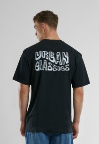 Fekete pamut póló, nagy, fehér grafikai felirattal "URBAN CLASSICS" a hátsó részén. A dizájn laza fazonnal és rövid ujjakkal rendelkezik.