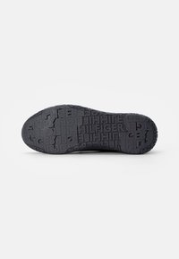 Semelle de chaussure noire avec motif répétitif embossé "HILFIGER" sur la surface de la bande de roulement, vue par le dessous sur fond blanc.
