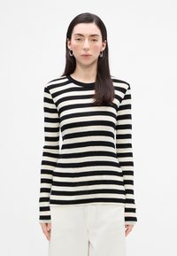 Camisa de rayas negra y crema de manga larga con cuello redondo, diseño ajustado y textura suave; combinada con pantalones de color claro.