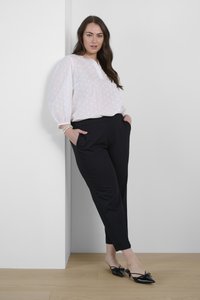 Blouse blanche à pois avec des manches bouffantes, assortie à un pantalon noir ajusté. Mules noires avec des nœuds sur un sol en bois contre un mur blanc.
