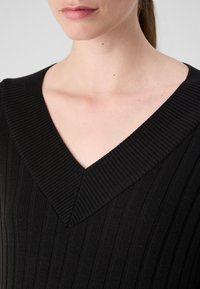 Melnā ribu adīta krekla ar dziļu V veida dekoltē, kas piedāvā vertikālas līnijas un teksturētu rakstu. Nav redzamu papildu detaļu vai rotājumu.