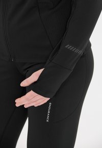 Sort atletisk jakke med ribbet tekstur på den ene side, tommelfingerhuller i ærmerne og et logo på den nederste del for "ENDURANCE."
