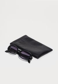 Óculos de sol pretos com lentes ovais, repousando sobre um estojo de couro preto. O estojo apresenta um logótipo estampado. As lentes são escuras e ligeiramente tintadas.