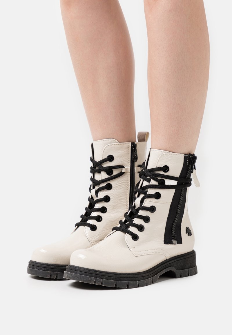 Bottes chevilles lacées couleur crème avec semelles noires et fermetures éclair latérales, portées sur des jambes nues sur fond blanc.