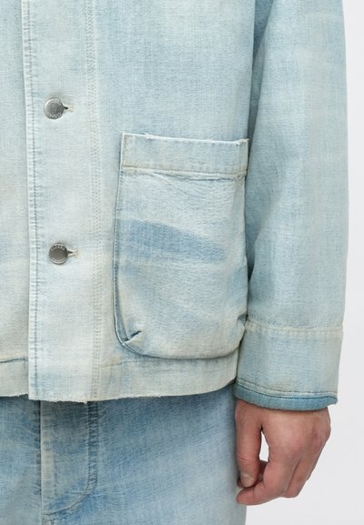 Ανοιχτό μπλε τζιν μπουφάν με τσέπη στο στήθος, ασημένιο κούμπωμα με κουμπί, και φθαρμένο τελείωμα με διακριτικές παραλλαγές υφής.