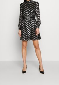 Robe noire avec des rayures diagonales argentées, un col haut, des manches longues et une taille cintrée. Fini avec une jupe évasée et des escarpins noirs élégants.