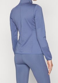 Lätt, figursydd, långärmad topp i en mjuk blått tyg med sömmar och texturerade paneler. Hög krage och platta sömmar.