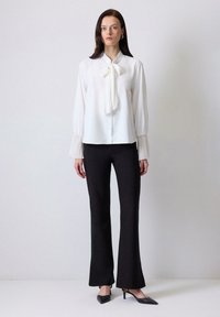 Blusa blanca con lazo y mangas con pliegues, combinada con pantalones negros de campana y tacones negros de punta. Fondo liso.