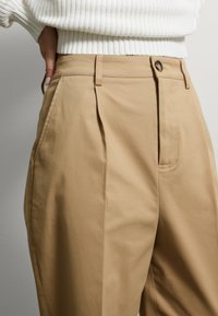 Pantalons plissés beiges en coton. Ils présentent une taille haute, des poches avant et une fermeture à un bouton, associés à un pull blanc texturé.