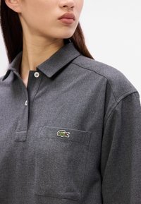 Polo gri din material moale, cu guler, nasturi și un buzunar pe pieptul stâng cu un logo brodat al unui crocodil verde.