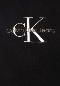 Tessuto in pile nero con testo ricamato bianco: "Calvin Klein Jeans" con logo "CK", che evidenzia una trama morbida e lussuosa.