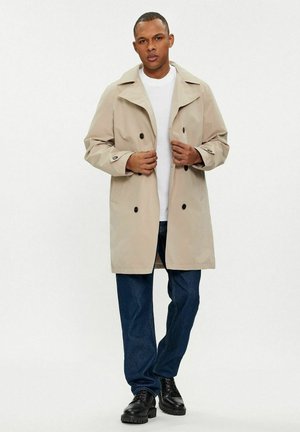 Trench coat beige con design doppiopetto, ampi rever e bottoni neri, indossato sopra una camicia bianca, abbinato a jeans scuri e scarpe nere.