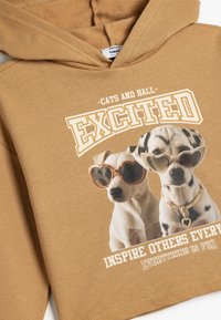 Sudadera marrón con un gráfico de dos perros usando gafas de sol. El texto arriba dice "EMOCIONADO" y debajo, "INSPIRAR A OTROS CADA DÍA". Material de algodón.