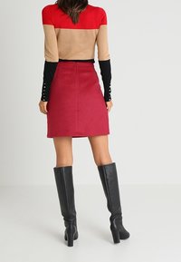 Femme portant un pull coloré, une jupe rouge et des bottes à talons montantes noires, se tenant devant un fond clair.