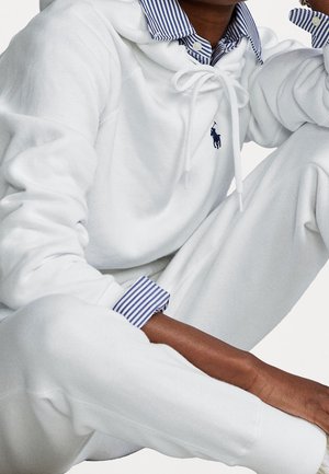 Personne portant un sweat à capuche blanc et un pantalon de survêtement avec des poignets et un col de chemise rayés bleus, assise avec un genou relevé, montrant le logo Polo Ralph Lauren.