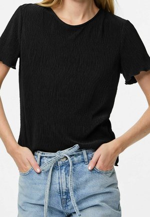 Kobieta w teksturowanej czarnej bluzce z krótkim rękawem, wpuszczonej w jasnoniebieskie dżinsy z wiązanym paskiem, ręce w kieszeniach.