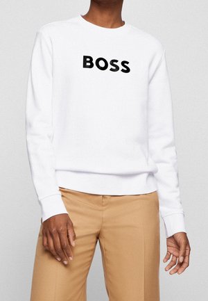 Osoba w białej bluzie z długim rękawem z wyraźnym czarnym napisem "BOSS", w połączeniu z beżowymi spodniami, pokazana od szyi do bioder na neutralnym tle.