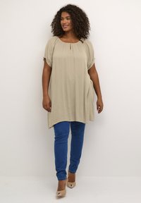 Tunika beige con collo tondo e dettaglio a chiave, abbinata a jeans skinny blu. Il modello indossa décolleté nude. Tessuto morbido e fluido.