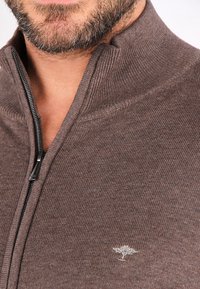 Brun zip-up genser med teksturert strikk, høy krage og en liten brodert trelogo på brystet. Glidelåsdetaljen er synlig.