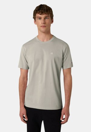 Giovane uomo in piedi, frontale, che indossa una T-shirt a maniche corte grigio chiaro con piccoli loghi sul petto e sulla manica.