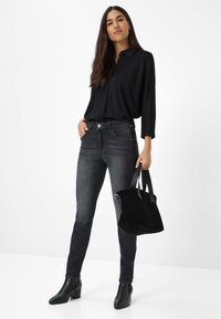 Femme debout en blouse noire, jeans skinny gris foncé, bottes noires à talons, tenant un sac à main en suède noir contre un fond blanc.