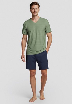 Man staat blootsvoets, draagt een groen T-shirt met korte mouwen en V-hals en marineblauwe shorts tegen een effen achtergrond.