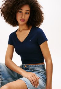 Νεαρή γυναίκα με σγουρά μαλλιά, που φοράει μπλε ναυτικό κοντομάνικο τοπ με V-λαιμόκοψη και ψηλόμεση τζιν φούστα, καθισμένη και κοιτάζοντας την κάμερα.