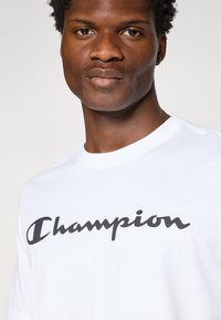 Weißes T-Shirt mit kurzen Ärmeln und schwarzem "Champion"-Logo in Schreibschrift auf der Brust. Einfaches Design, aus Baumwolle, runder Halsausschnitt.