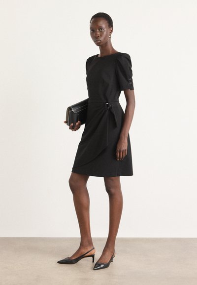 Robe noire à manches courtes avec un décolleté carré, épaules froncées, drapés sur les côtés et une ceinture. Portée avec des chaussures à talons ouverts noires et une pochette.
