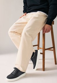 Pantalon en coton beige clair à coupe décontractée, associé à des baskets foncées avec des semelles blanches, assis sur un tabouret en bois sur un fond neutre.