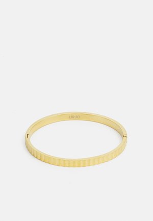 Kultainen bangle-rannekoru, jossa on uurrettu pinta, ja joka sisältää sileän, kiillotetun sisäpuolen sekä pienen logopainatuksen.