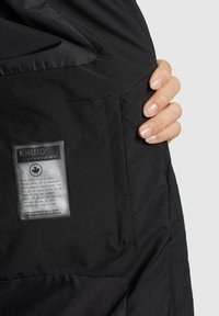 Veste noire avec un extérieur texturé, une étiquette brillante avec du texte et une poche. Le design comprend un tissu lisse et des détails de couture visibles.