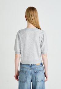 Vrouw met lang blond haar, gekleed in een lichtgrijze trui met korte mouwen en hoge taille blauwe jeans, staande met haar gezicht naar weg tegen een witte achtergrond.