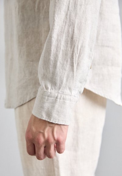 Chemise en lin beige à manches longues avec une coupe décontractée, poignets boutonnés et un tissu texturé. Main serrée près de l'ourlet de la chemise.