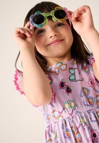 Barevné sluneční brýle ve tvaru květiny držené nad fialovými šaty s výraznými vzory slunečních brýlí, hvězd a ovoce s růžovou lemovkou z bambulí.