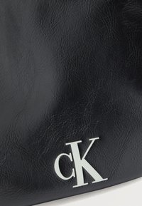 Κοντινό πλάνο μαύρης υφασμάτινης επιφάνειας με λευκό λογότυπο "cK" στην κάτω δεξιά γωνία, που υποδηλώνει την επωνυμία Calvin Klein.