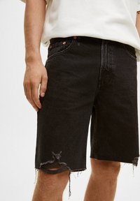 Shorts de mezclilla negra con dobladillo deshilachado, detalles desgastados en la pierna, diseño de cinco bolsillos y acentos de hardware en tono cobre.