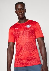 Crvena majica kratkih rukava s teksturiranim uzorkom, Puma logotipom i ekipnim grbom na prsima. Izrađena od prozračnog materijala.