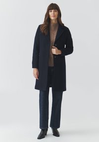 Navy wool blend coat with a notched collar, two buttons, and side pockets; worn over a beige cable-knit sweater and dark jeans.