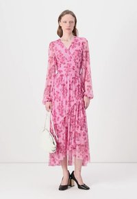 Robe portefeuille florale rose avec de longues manches transparentes, un ourlet à volants et une ceinture à la taille. Portée avec des chaussures à talons bloc noires et un sac à main blanc.