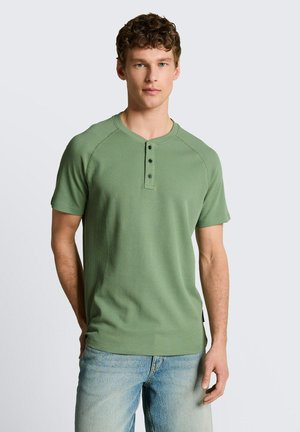 Jonge man met krullend haar die een saliegroene short-sleeve henley draagt en lichtblauwe jeans, staand tegen een effen witte achtergrond.