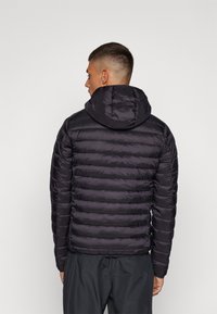 Giacca nera imbottita con cappuccio, caratterizzata da un design trapuntato e polsini elasticizzati, vista da dietro. Materiale leggero con una texture liscia.
