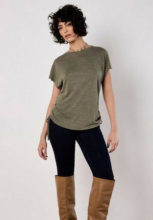 Haut olive vert à manches courtes avec une finition texturée, doté de liens sur les côtés. Associé à un jean bleu foncé et des bottes en cuir beige jusqu'aux genoux.