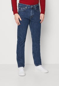 Jeans de mezclilla azul oscuro con corte recto, cinco bolsillos y costuras en contraste; llevados con zapatillas blancas y una camiseta de manga larga roja.
