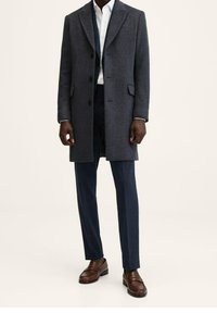Dunkelgrauer Wollmantel mit taillierter Passform, einreihigem Frontverschluss und Reverskragen, kombiniert mit marineblauen Karohosen und braunen Lederschuhen.