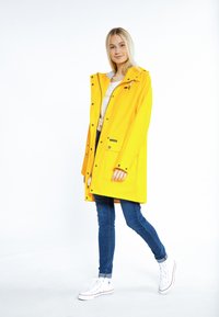 Gelbe wasserdichte Jacke mit Kapuze, Frontreißverschluss und Druckknöpfen, zusammen mit blauen Jeans und weißen Sneakern in einer hellen Umgebung präsentiert.