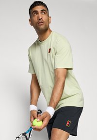 Light green cotton T-shirt with short sleeves, featuring a small multicolored logo patch. Paired with dark shorts and gripping a tennis ball.