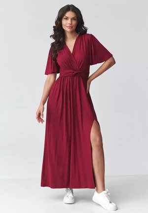 Kobieta ubrana w długą burgundową sukienkę kopertową z krótkimi rękawami i rozcięciem z boku, zestawioną z białymi trampkami, stojąca na jednolitym tle.