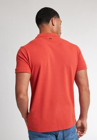 Červené bavlněné polo triko s krátkým rukávem, límcem a malým logem na zadní straně blízko krku. Volný střih a hladká textura.